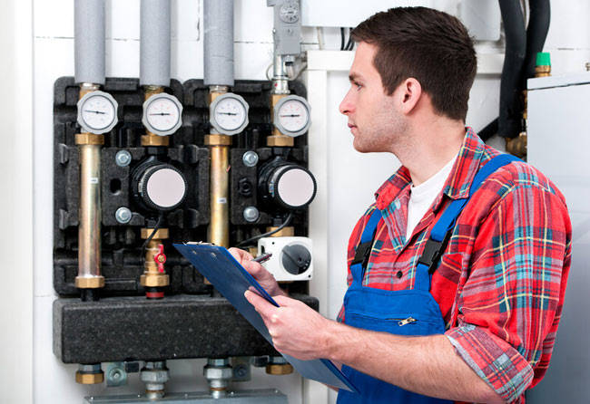 An HVAC technician checks gauges and pipes on a boiler system while taking notes on a clipboard.